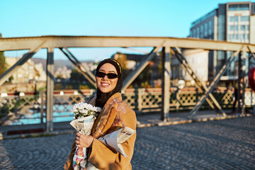 A hijab woman in stylish sunglasses and an elegant French outfit, walking through the city at sunset, carrying a bouquet, bread, and newspaper, radiating a sense of cultural charm and serenity