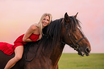 Obraz premium A young woman in a red dress walks with a horse in a field at sunset