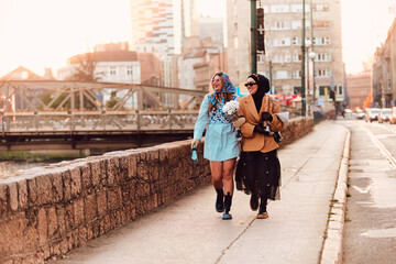 Couple woman one wearing a hijab and a modern yet traditional dress, and the other in a blue dress and scarf, walking together through the city at sunset. One carries a bouquet and bread, while the