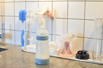 A baby milk bottle next to a baby bottle dryer and a blue baby bottle brush.
