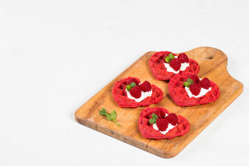 Red heart-shaped waffles with cream cheese, raspberries and micro greenery. Waffles with the addition of beet juice. On a serving wooden board. White background. Valentine's Day. Copy space