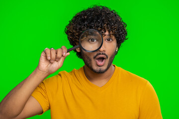 Investigator researcher scientist man holding magnifying glass near face, looking into camera with big zoomed funny eyes, searching, analysing. Handsome indian guy isolated on chroma key background