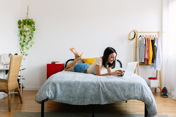 Happy young woman using laptop lying on bed at home. Millennial teenage girl working or studying...