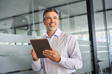 Happy middle aged business man ceo standing in office using digital tablet. Smiling mature...