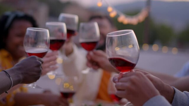 Group of young multiracial friends together celebrating birthday by toasting with wine on rooftop in evening. Happy and cheerful people dining gathered sitting at table outdoor. Social relationships.