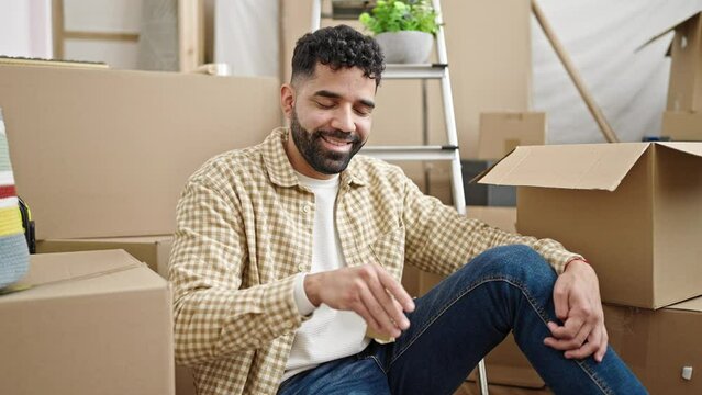 Young hispanic man holding keys sitting on floor at new home