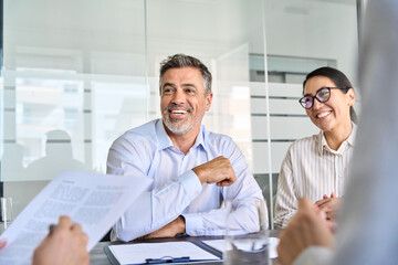 Happy diverse business people international corporate executive team talking at group meeting. Smiling older Latin male manager working with colleagues collaborating at boardroom table.
