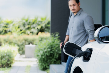 Focus electric car charging at home charging station with blurred progressive man walking in the...