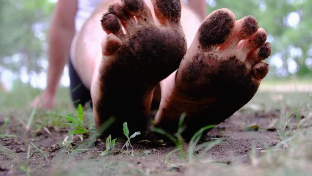 Showing to camera bare soles full of mud after waling through field or farm land after rain. Man wearing white shirt and only black briefs.