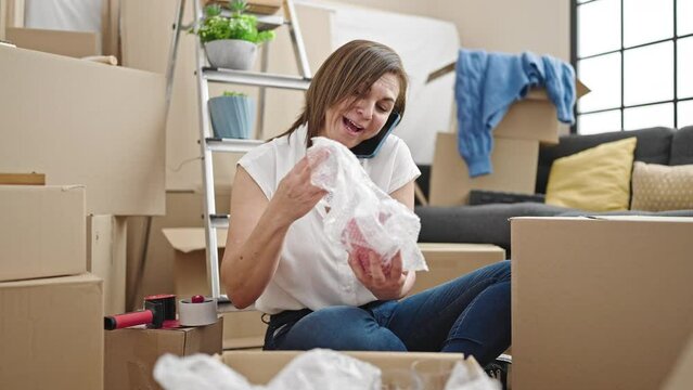 Middle age hispanic woman unpacking cardboard box speaking on the phone at new home