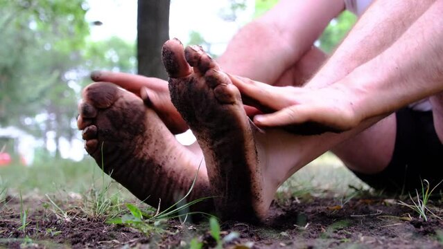 Close up while rubbing bare underfoot or soles with mud getting them dirty while sitting on the ground. Man wearing white shirt and only black briefs.