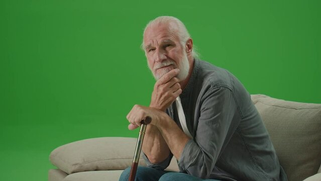 Green Screen. Portrait of a Sad Old Man with a Stick Is Sitting On A Sofa. An Elderly Man Leaning His Head On A Stick and Rubs His Gray Beard. Mental Health for Seniors.
