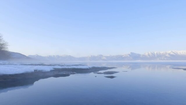 Hot spring steam drifting over icy shore and calm water of caldera lake at dawn in winter