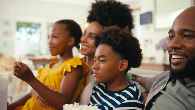 Family at home sitting on sofa together streaming show or movie to TV with daughter throwing popcorn at father who catches it in mouth and eats it - shot in slow motion 