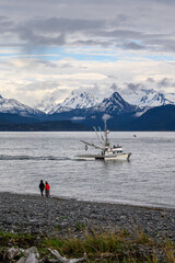Fototapeta na wymiar Fishing boat heading out of Katchemak Bay in the morning to go fishing, people walking on beach, mountain range in the background, Homer, AK 