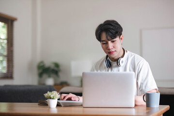 A focused young Asian male college student or freelancer using his digital tablet at a table in his living room. - Powered by Adobe