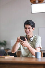 A cheerful and smiling young Asian man in comfy clothes enjoys playing mobile games on his phone...