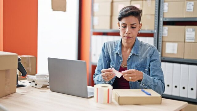 Young beautiful hispanic woman ecommerce business worker using laptop putting sticker on package at office