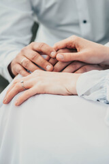 a young couple in light clothes sit and hold hands, there is a ring on her finger