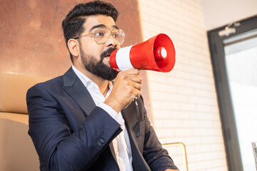 Young indian businessman in suit and glasses shouting in megaphone. Male entrepreneur screaming in loudspeaker.