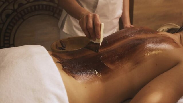attractive fair-haired woman gets pleasure for receiving chocolate body mask. Slow motion. Chocolate body wrapping. SPA. Young beautiful girl lies on massage table.