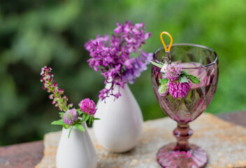 A glass with a drink and flowers in a white vase on the terrace against the backdrop of a green garden