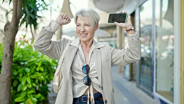 Middle age blonde woman smiling confident using smartphone with winner expression at street