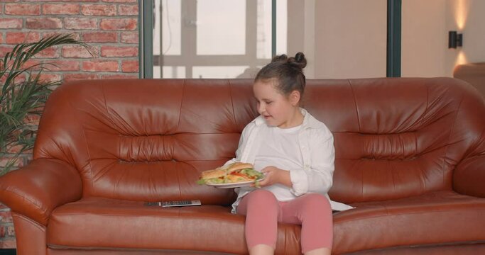happy cute plump kid walking with a plate of burges at home, sitting on couch, going to have snack as she is hungry Slow motion