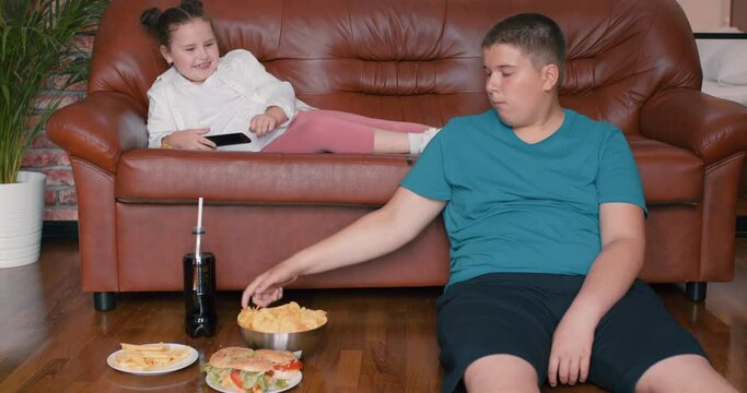 Overweight fat boy sitting on floor, plump girl lying on sofs they eat fast food, burgers , chips, drink coca cola, watch TV, cartoon, film, movie Slow motion lifestyle Leisure