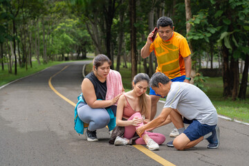 A young woman in fitness clothes with knee pain grabbing her knee with both hands while sitting on the middle of a running track at a local park with three other runners gather around trying to help