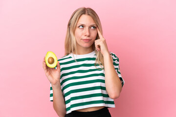 Young caucasian woman holding an avocado isolated on pink background having doubts and thinking