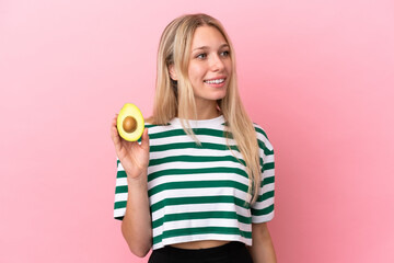 Young caucasian woman holding an avocado isolated on pink background looking to the side and smiling