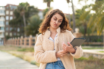 Young caucasian woman holding a tablet at outdoors with sad expression