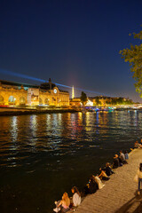 夜のセーヌ川沿いでくつろぐ人々とエッフェル塔などが輝くパリの夜景