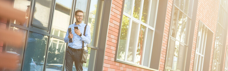 Business talk. Young confident caucasian businessman wearing blue shirt and eyeglasses holding his laptop and talking by phone while walking outdoors