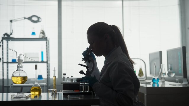 Medical scientific laboratory. Side view of a dark silhouette of a female scientist looking under a microscope, doing an analysis of a test sample.