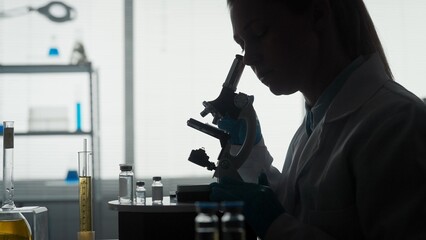 Side view of a dark silhouette of a female scientist looking under a microscope, doing an analysis of a test sample close up. Ambitious biotechnology specialist, working with advanced equipment.