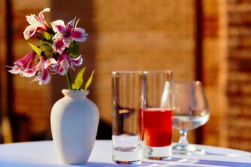 A vase with a flower and glasses on a table in the courtyard before sunset
