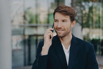 Handsome exec hears vital info with fast mobile, dressed formal, poses near blurred background, uses roaming.