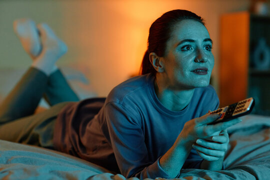 Portrait of adult woman lying on bed at home and watching TV in dark