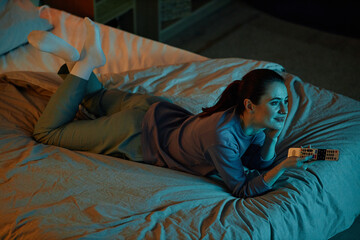 Fototapeta na wymiar High angle portrait of adult woman watching TV in dark while lying on bed at home, copy space