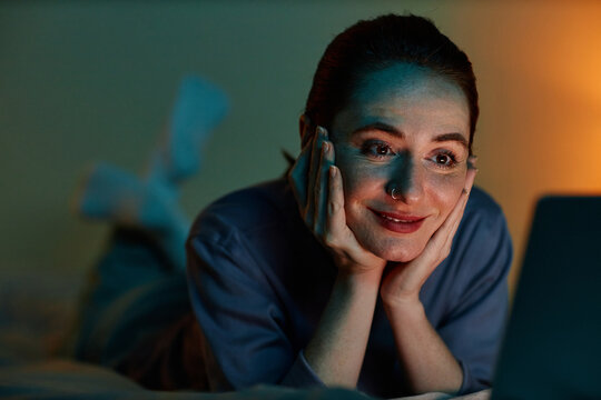 Closeup portrait of young woman binge watching movies at night while lying on bed with laptop