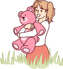 Happy little girl with teddy bear in hands stands in meadow among tall grass and looks at screen