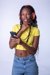 Mujer hermosa joven de color que usa el teléfono inteligente pensativa en una fondo blanco. Chica estudiante sonriente enviando mensajes de texto en el teléfono móvil con los auriculares puestos. - obrazy, fototapety, plakaty