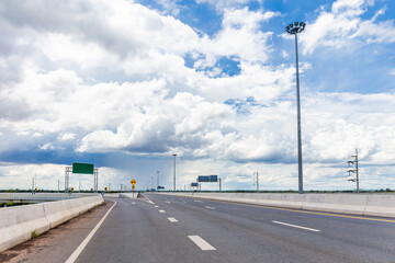 Heavy concrete road with lampposts, spotlights and traffic signs on a cloudy blue sky day.