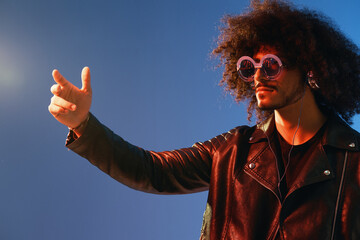 Portrait of a stylish man with curly hair with glasses and headphones on a blue background multinational, colored light, black leather jacket trend, modern concept.