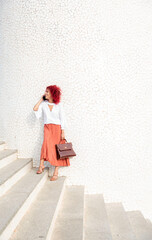 elegant business woman with red afro hair on white stairs holding handbag in hand leaning against white wall talking on smartphone