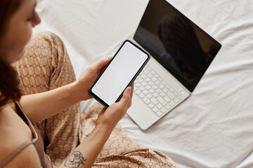 Minimal top view at young woman using laptop and smartphone with blank white screen on bed, copy space