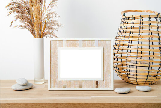 Minimal natural wood home decor with white empty picture frame, dry reed in vase and round wood lantern with candle burning. Home interior background.