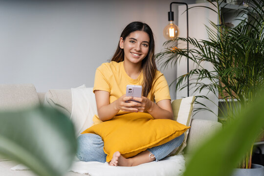 Cheerful female student with beautiful smile on cosy sofa at home holding a phone looking in the camera